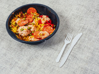 Spanish paella dish in black round plastic tray on light color table cloth and plastic forks and knife. Popular food with rice and sea products and meat. Fast food product.