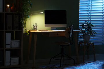 Comfortable workplace with computer and lamp on wooden desk at home office in evening