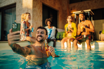 Man taking selfie while drinking beer in swimming pool with friends