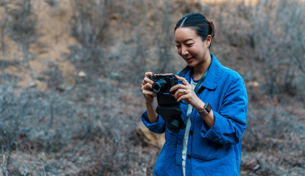 Young asian woman photographer wearing blue jacket, smiling while reviewing photos she just took with her digital camera in a natural outdoor setting
