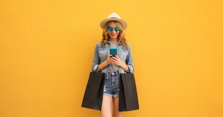 Beautiful happy young woman holding phone with black shopping bag, summer hat on yellow background