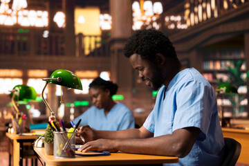 Black med student in scrubs does research and takes notes for his thesis, working hard at the...