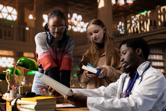 Diverse group of learners engages in a discussion with physician tutor, utilizing textbooks and class notes. Using university library resources to expand their medical knowledge on medicine research.