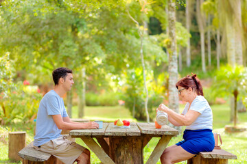 Naklejka premium Mother and adult son enjoying a relaxing coffee break at a wooden picnic table in a green park. Concept of family love, bonding and Mother's Day