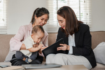 Fototapeta premium Two women and a baby in a home setting, discussing business with a tablet, showcasing work-life balance and modern family dynamics.