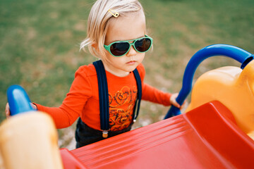 Fototapeta premium Little girl in sunglasses climbs a slide holding onto the handrails