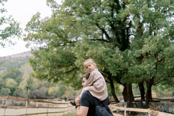 Little girl looks around while sitting on her dad shoulders as he walks through a green park. Back view