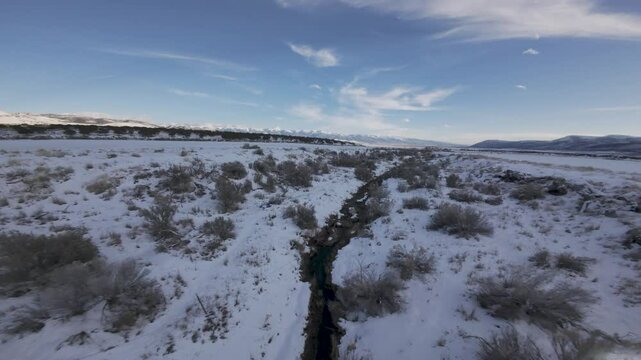 Aerial-First Person View-FPV-Flying low and fast over a small snow lined stream with tall sagebrush under a blue winter sky with high clouds