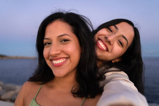 Two happy hispanic women taking a selfie by the sea at sunset