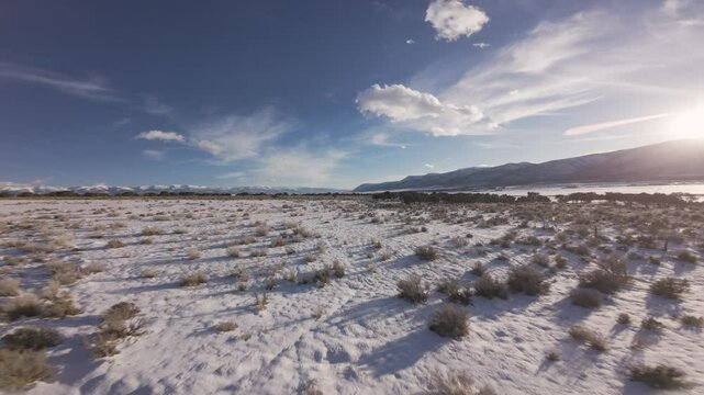 Aerial-FPV-First person view- Flying low over snowy sagebrush flat-Low sun casting long shadows with sun glare peeking over the mountain under dark blue sky under high wispy clouds in winter scene