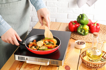 Woman frying vegetables in pan on stove at wooden table indoors, closeup