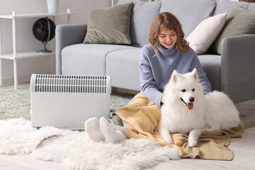 Young woman with plaid and Samoyed dog warming near radiator on floor at home