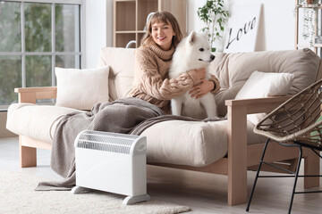 Young woman with blanket and Samoyed dog on sofa warming near radiator at home