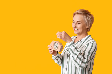 Beautiful mature woman putting coins into wooden piggy bank on yellow background