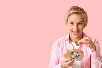 Beautiful mature woman putting coins into wooden piggy bank on pink background