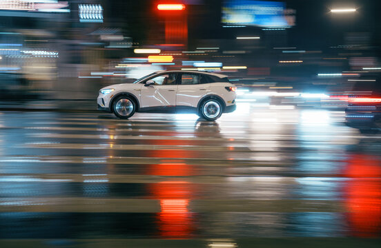 White car driving through busy wet street intersection on a rainy night. Long exposure. Bright colorful city lights blurred in background.