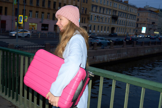 Young woman with pink suitcase walks along river in city