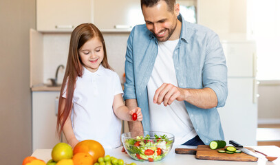 Happy Little Girl Cooking With Daddy Preparing Vegetable Salad For Dinner Seasoning Dish Standing In Modern Kitchen At Home. Father And Daughter Enjoying Food Preparation On Weekend