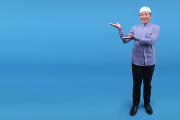 Full Length Shot Of Young Asian Muslim Man Smiling And Presenting Aside To Ads Space Over Blue...