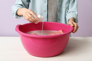 Woman adding detergent to clothes in plastic basin on table against lilac background