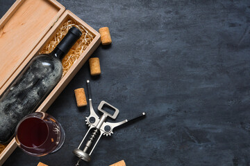 Box with bottle of wine, glass, corks and opener on black grunge background