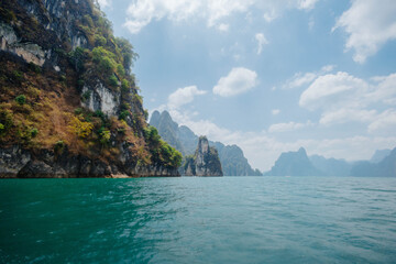 Obraz premium Rocky cliffs rise dramatically from turquoise waters, surrounded by lush greenery. calm lake reflects blue skies with fluffy clouds, creating a serene atmosphere on a clear day