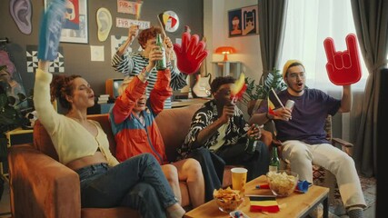 Group of excited young friends sitting on couch by table full of snacks and drinks, cheering for favorite team, waving Belgium flags and foam fingers, watching sports game on TV in stylish living room
