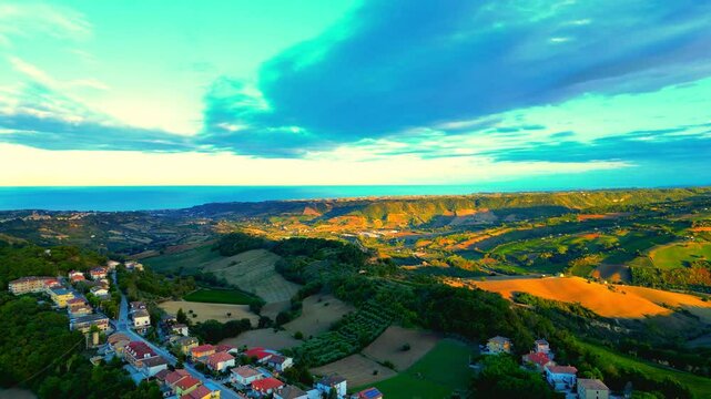 Gorgeous aerial push-in shot from Montefiore dell'Aso (AP), the vast mirroring azure expanses of the Adriatic Sea as a backdrop for balmy Marche hillscapes permeated by reddish light, cloudy blue sky