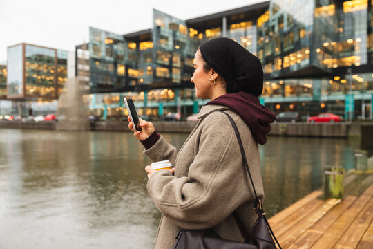 Student in niqab by city river enjoying coffee and photography