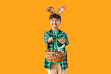 Cute boy in bunny ears holding wicker basket with sweet chocolate eggs and toy rabbit on yellow background. Easter celebration