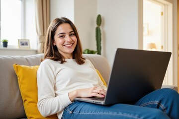 Fototapeta premium Cheerful Young Caucasian Woman Using Laptop While Sitting Comfortably on Sofa in Stylish Living Room with Natural Light and Cactus Decoration