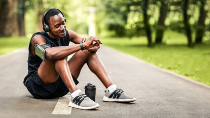 Athletic black man sitting on park way, checking on his smartwatch, measuring pulse, empty space