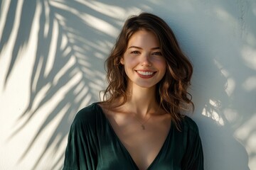 Cheerful young attractive woman in stylish dark-green top enjoys sunny weather, poses near white wall with palm leaf shadow and smiles