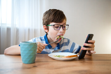 Boy with glasses watches a mobile phone while eating in a modern kitchen. The concept of health risks for screen time