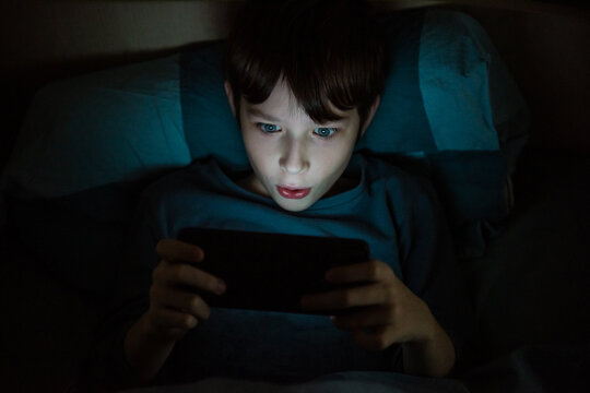 A teenage boy at night under a blanket with a phone. Children who are addicted to gadgets. Violation of mental health due to gadgets. Visual impairment.