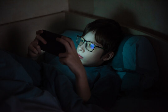 Teenage boy with glasses at night under a blanket with a smartphone. Children who are addicted to gadgets. Mental health problems caused by gadgets. Visual impairment.