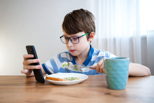 Cute boy with glasses is watching a mobile phone while eating in a modern kitchen. The concept of performance risks for screen time. Risk to vision. Dependence on gadgets