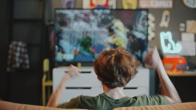 Young man sitting on couch, excitedly watching soccer match on TV and enthusiastically cheering for favorite team in living room with modern decor. Back view