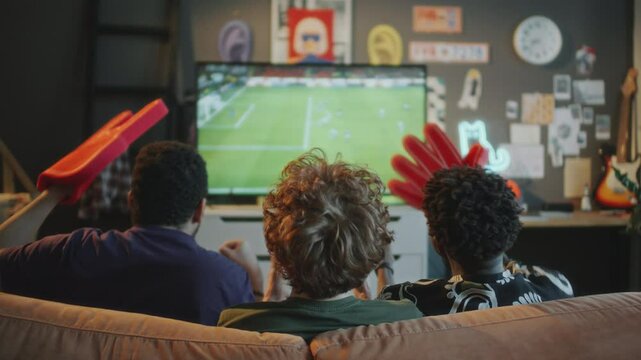 Three male friends sitting on sofa in living room, celebrating goal and high fiving each other with foam fingers as watching soccer match on TV at home. View from behind