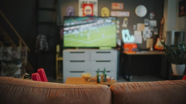 Empty living room with couch in foreground and TV displaying soccer match, coffee table with snacks and drinks in defocused background