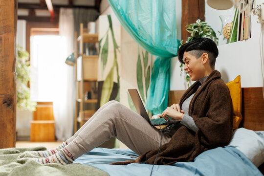 Cheerful woman in bedroom using laptop computer in cozy apartment