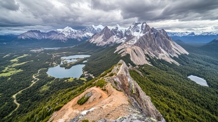 Jagged mountain peak overlooks lush valleys, lakes, and snowy mountains under cloudy skies