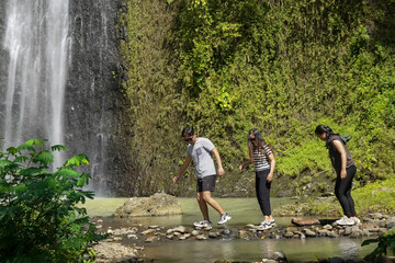 group of young asian indonesian man and women walking and crossing small stream with waterfall on the background