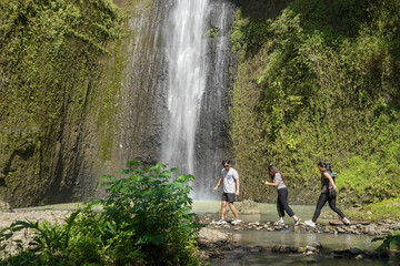 group of young asian indonesian man and women walking and crossing small stream with waterfall on the background
