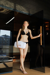 Fototapeta premium Smiling woman holding towel and standing near sauna in spa complex