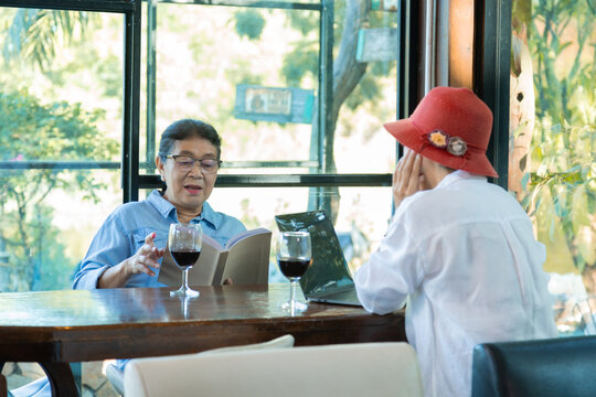 Two Asian lifestyle elderly females after retirement  enjoy leisurely afternoons in cafe,  focused on reading book, using laptop for working, enjoying social time together in charming settings.