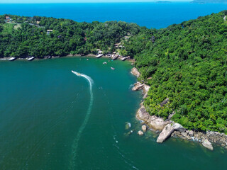 Fototapeta premium A stunning aerial shot captures the lush green coastline and deep blue waters of Praia do Cedro. A speedboat leaves a white trail as it glides through the calm sea.