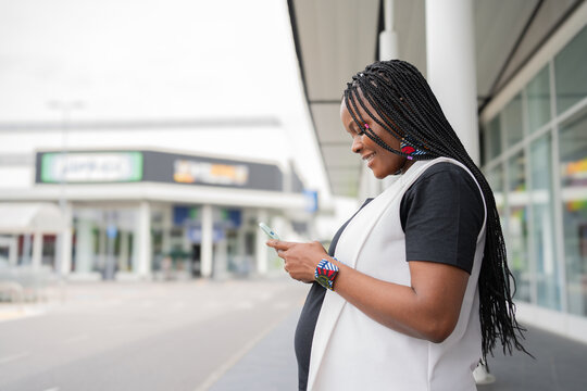 Donna incinta di origine africana con capelli intrecciati sorride mentre usa lo smartphone all&rsquo;esterno di un centro commerciale. Indossa un outfit casual con accessori colorati, trasmettendo felicit&agrave; 