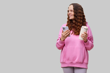 A woman with wavy hair stands in a relaxed pose, holding a coffee cup in one hand and a smartphone...