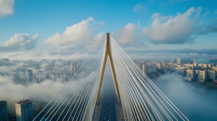 Obraz premium Aerial view of a cable stayed bridge piercing a low lying cloud layer above a city skyline on a bright day. The bridge's structure is sharp against the soft clouds and cityscape.